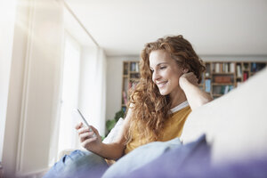 Lächelnde Frau zu Hause auf der Couch sitzend und auf das Handy schauend - RBF003128