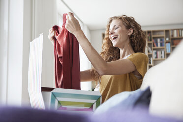 Happy woman at home sitting on couch unpacking parcel with garment - RBF003085