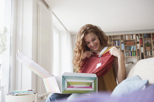 Smiling woman at home sitting on couch unpacking parcel with garment - RBF003087