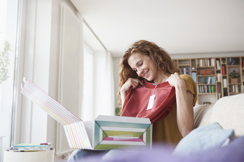 Lächelnde Frau zu Hause auf der Couch sitzend beim Auspacken eines Pakets mit Kleidungsstücken, lizenzfreies Stockfoto