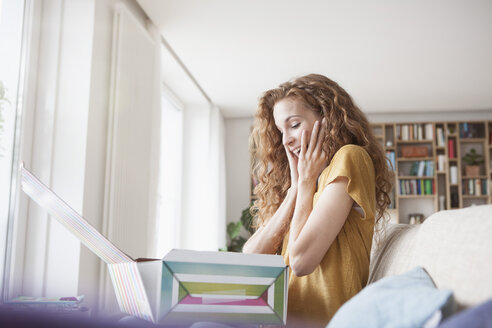 Amazed woman at home sitting on couch unpacking gift box - RBF003089