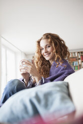 Lächelnde Frau zu Hause auf der Couch sitzend und eine Tasse haltend - RBF003092