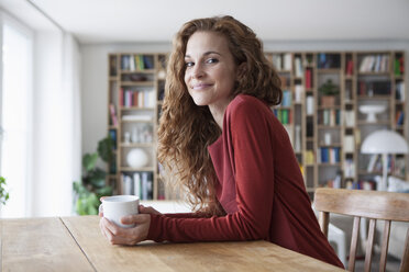 Smiling woman at home sitting at wooden table with cup - RBF003116