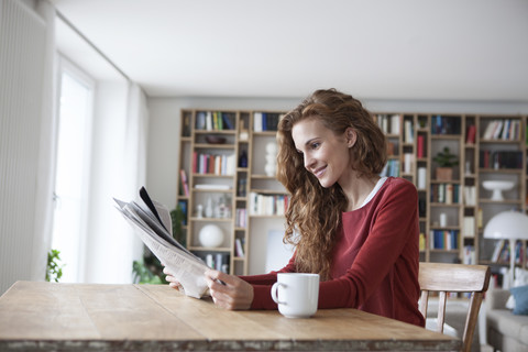 Lächelnde Frau sitzt zu Hause am Holztisch mit Tasse und liest Zeitung, lizenzfreies Stockfoto