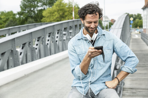Lächelnder Mann auf einer Brücke schaut auf sein Handy - FMKF001879