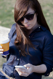 Porträt einer Frau mit Sonnenbrille auf einer Parkbank sitzend, mit Smartphone und Kaffee zum Mitnehmen - HCF000147