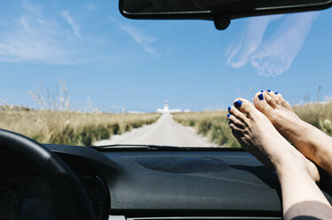 Spain, Menorca, feet on the dashboard, driving on empty road on vacations with a lighthouse in the background - JRFF000004