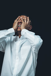 Laughing young man wearing white shirt with hands on his face in front of black background - CHAF001368