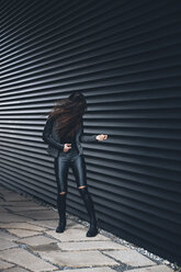 Schwarz gekleidete junge Frau, die vor einer schwarzen Fassade steht und Luftgitarre spielt - CHAF001379