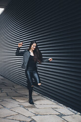 Schwarz gekleidete junge Frau hüpft auf einem Bein vor einer schwarzen Fassade - CHAF001380