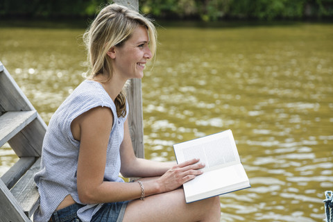 Lächelnde junge Frau beim Lesen eines Buches am Wasser, lizenzfreies Stockfoto
