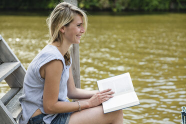 Lächelnde junge Frau beim Lesen eines Buches am Wasser - FMKF001909
