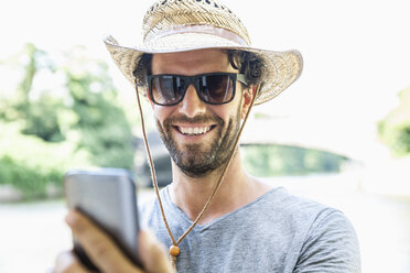 Lächelnder Mann mit Strohhut und Sonnenbrille, der ein Mobiltelefon hält - FMKF001915