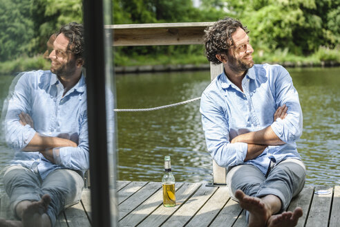 Smiling man sitting on platform at the waterside with beer bottle - FMKF001965