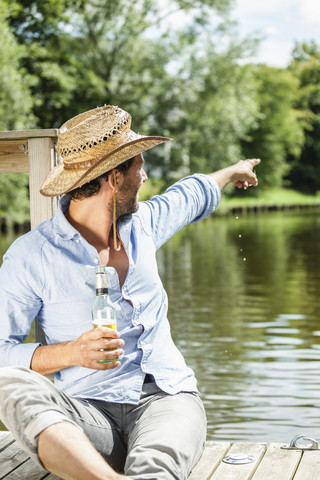 Mann sitzt auf einer Plattform am Wasser mit einer Bierflasche und zeigt mit dem Finger darauf, lizenzfreies Stockfoto