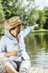 Mann sitzt auf einer Plattform am Wasser mit einer Bierflasche und zeigt mit dem Finger darauf - FMKF001966