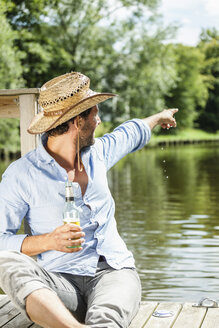 Mann sitzt auf einer Plattform am Wasser mit einer Bierflasche und zeigt mit dem Finger darauf - FMKF001966