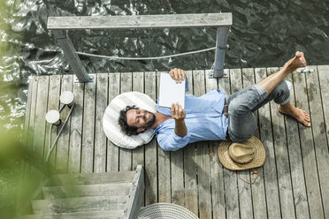 Man lying on platform at the waterside holding digital tablet - FMKF001969