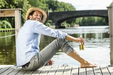 Lächelnder Mann, der mit einer Bierflasche auf einer Plattform am Wasser sitzt - FMKF001983