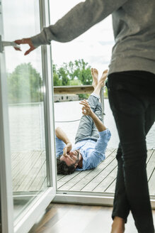 Man lying on wooden deck of a house boat with woman opening balcony door - FMKF001975