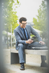 Geschäftsmann sitzt auf einer Bank und schaut auf sein Smartphone - UUF005371