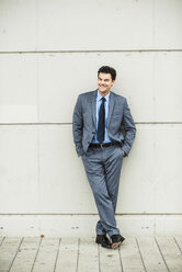 Porträt eines lächelnden Geschäftsmannes im grauen Anzug, der an einer Betonwand lehnt - UUF005373