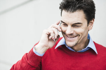 Portrait of smiling businessman wearing red pullover telephoning with smartphone - UUF005381