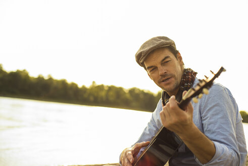 Portrait of man playing guitar at riverside in the evening - UUF005384