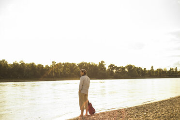 Mann mit Gitarre am abendlichen Flussufer stehend - UUF005386
