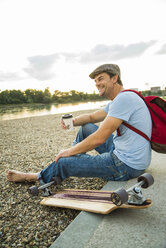 Lächelnder Mann am steinigen Strand mit Skateboard und Coffee to go am Abend - UUF005399