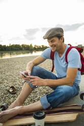 Lächelnder Mann am steinigen Strand mit Skateboard und Smartphone am Abend - UUF005400