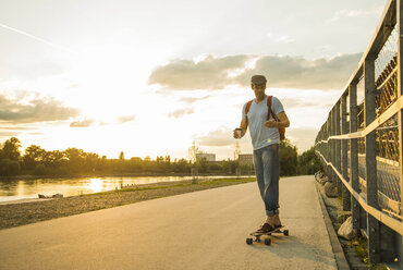 Man with coffee to go and smartphone standing on skateboard in the evening twilight - UUF005404