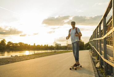 Mann mit Coffee to go und Smartphone auf Skateboard in der Abenddämmerung stehend - UUF005404
