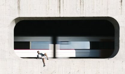 Spanien, Tarragona, entspannter junger Mann in Nische einer Betonwand mit fahrendem Zug im Hintergrund - JRFF000008