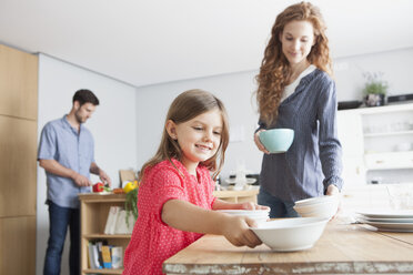 Lächelndes kleines Mädchen deckt den Tisch in der Küche mit ihren Eltern im Hintergrund - RBF003367