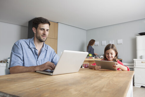 Mann und seine kleine Tochter sitzen am Küchentisch und benutzen einen Laptop und ein digitales Tablet - RBF003372