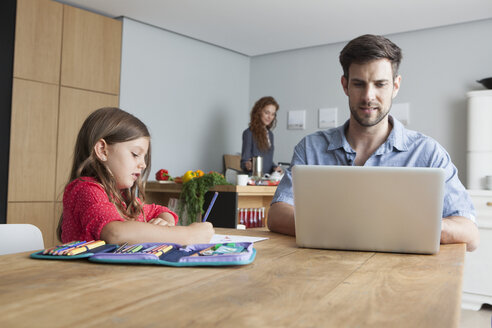 Mann sitzt am Küchentisch mit Laptop, während seine kleine Tochter malt - RBF003373