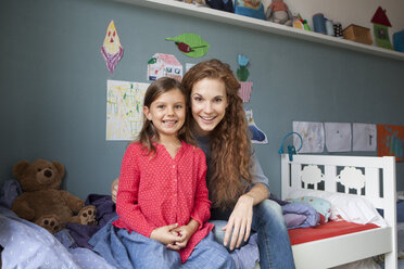 Porträt von Mutter und kleiner Tochter, die nebeneinander auf einem Bett im Kinderzimmer sitzen - RBF003387