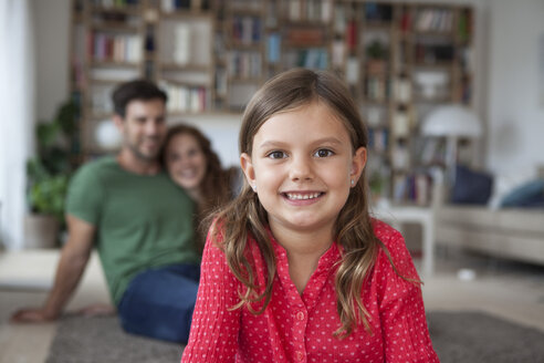 Porträt eines lächelnden kleinen Mädchens und ihrer Eltern im Hintergrund im Wohnzimmer - RBF003389