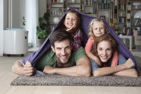 Porträt eines Paares mit ihren kleinen Töchtern, die zusammen auf dem Boden des Wohnzimmers liegen, zugedeckt mit einer Decke, lizenzfreies Stockfoto