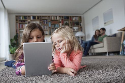 Zwei kleine Schwestern liegen auf dem Boden im Wohnzimmer mit digitalem Tablet, während ihre Eltern im Hintergrund sitzen - RBF003397