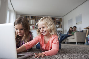 Two little sisters lying on the floor at living room with laptop while her parents sitting in the background - RBF003398