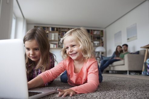 Zwei kleine Schwestern liegen auf dem Boden im Wohnzimmer mit Laptop, während ihre Eltern im Hintergrund sitzen - RBF003398