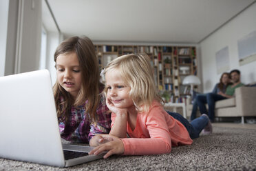 Zwei kleine Schwestern liegen auf dem Boden im Wohnzimmer mit Laptop, während ihre Eltern im Hintergrund sitzen - RBF003399