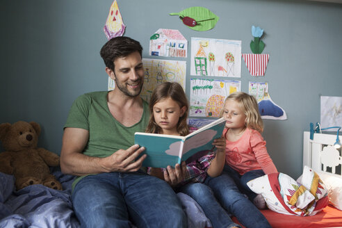 Vater und seine kleinen Töchter sitzen zusammen auf dem Bett im Kinderzimmer und lesen ein Buch - RBF003400
