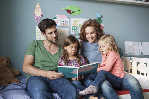 Ehepaar und kleine Töchter sitzen zusammen auf dem Bett im Kinderzimmer und lesen ein Buch - RBF003401