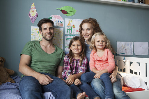 Family picture of couple with her little daughters sitting together on bed in children's room - RBF003402