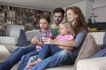 Couple sitting with their with two little daughters on couch in the living room looking at digital tablet - RBF003417