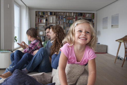 Porträt eines lächelnden kleinen Mädchens auf der Rückenlehne einer Couch mit ihrer Familie im Hintergrund - RBF003419