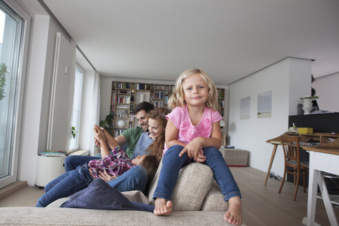 Porträt eines kleinen Mädchens, das auf der Rückenlehne einer Couch sitzt, mit ihrer Familie im Hintergrund - RBF003420